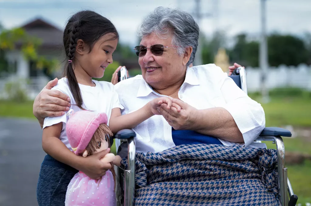 A elderly grandmother sitting in a wheelchair is smiling and holding hands with her granddaughter